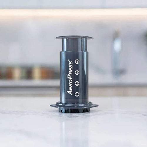 Cafetera AeroPress negra sobre mesa blanca con fondo desenfocado de cocina moderna