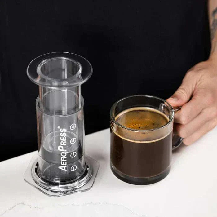 cafetera AeroPress Clear transparente al lado de taza de café espresso