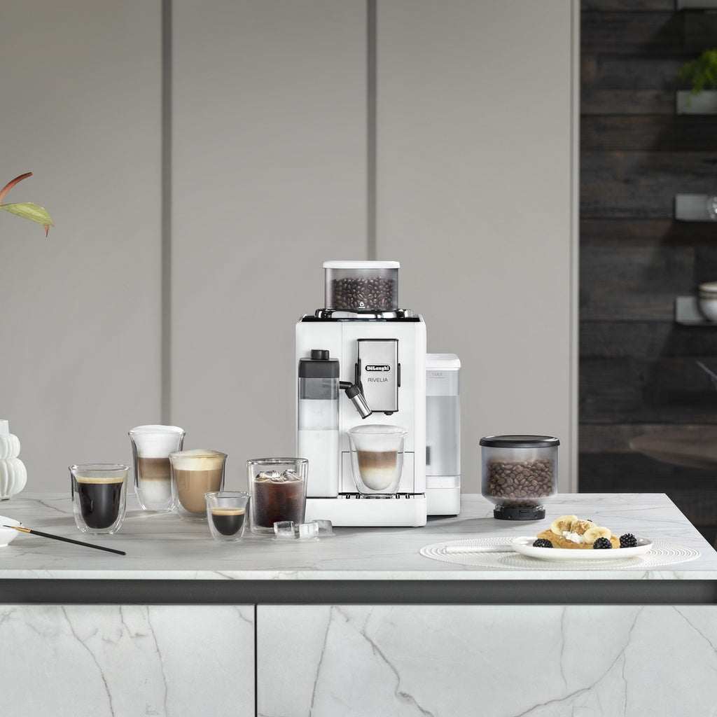 White Delonghi Rivelia super-automatic coffee machine with 1kg Explora Convención, featuring a touch screen and LatteCrema Hot technology, on a kitchen counter with various coffee drinks.